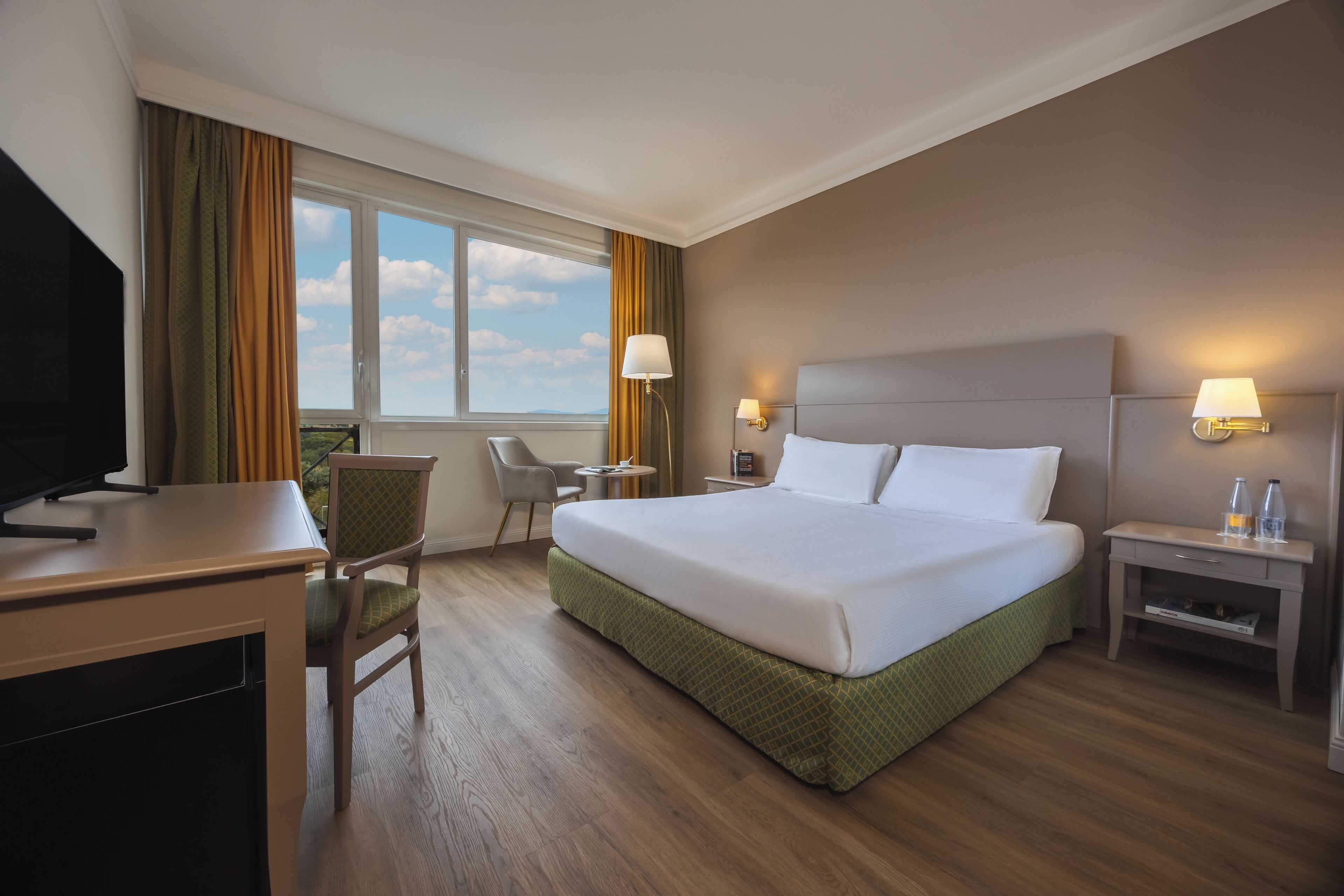 A hotel room with a bed, desk, chair, bedside tables, lamps, and a window with curtains.