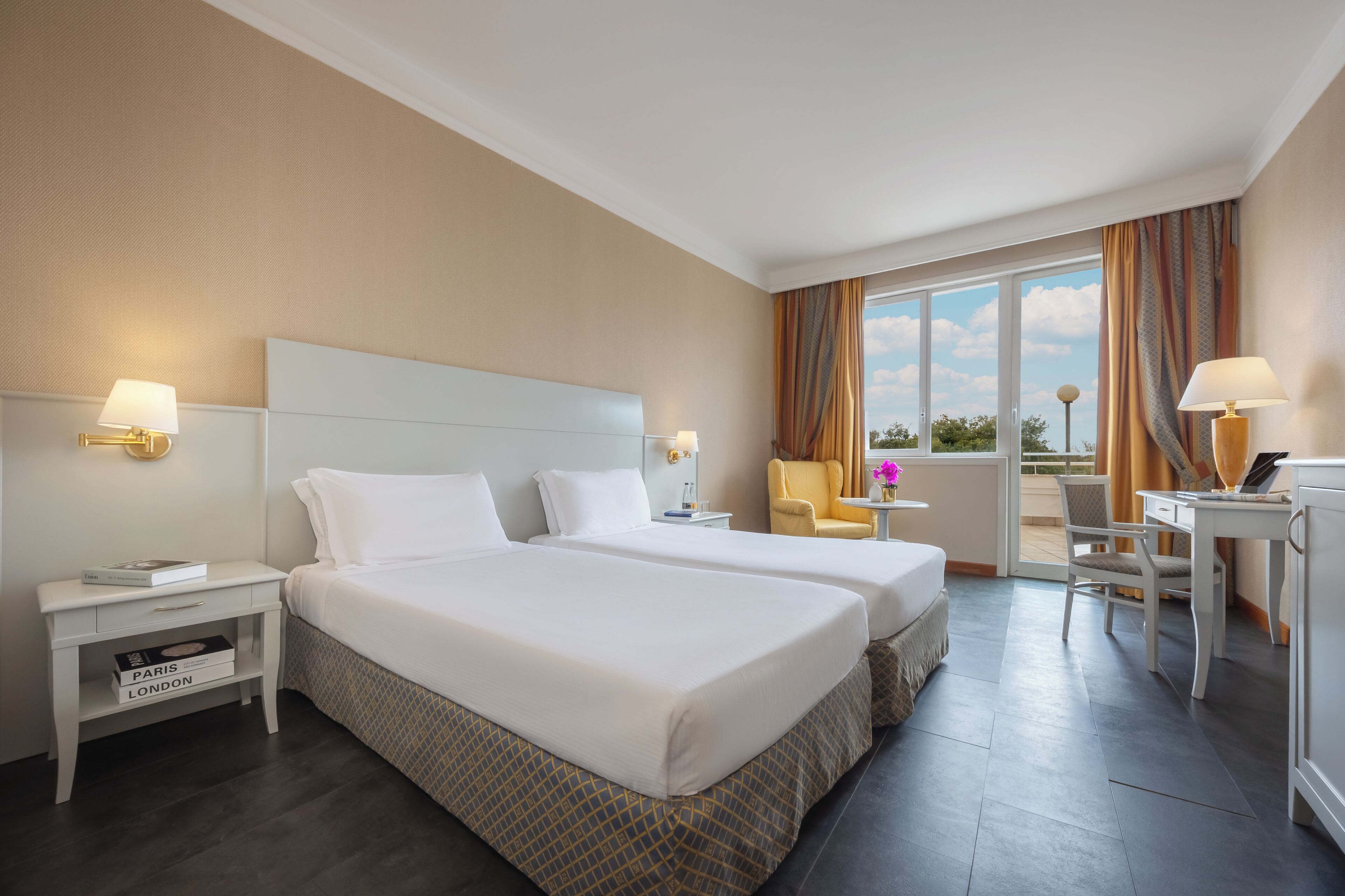 A hotel room with two beds, a desk, a chair, a nightstand, and a view of the outdoors.