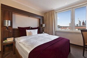 A hotel room with a large bed, two bedside lamps, a nightstand, a chair, and a window with a city view. - Motel One Frankfurt-Riverside (Frankfurt)