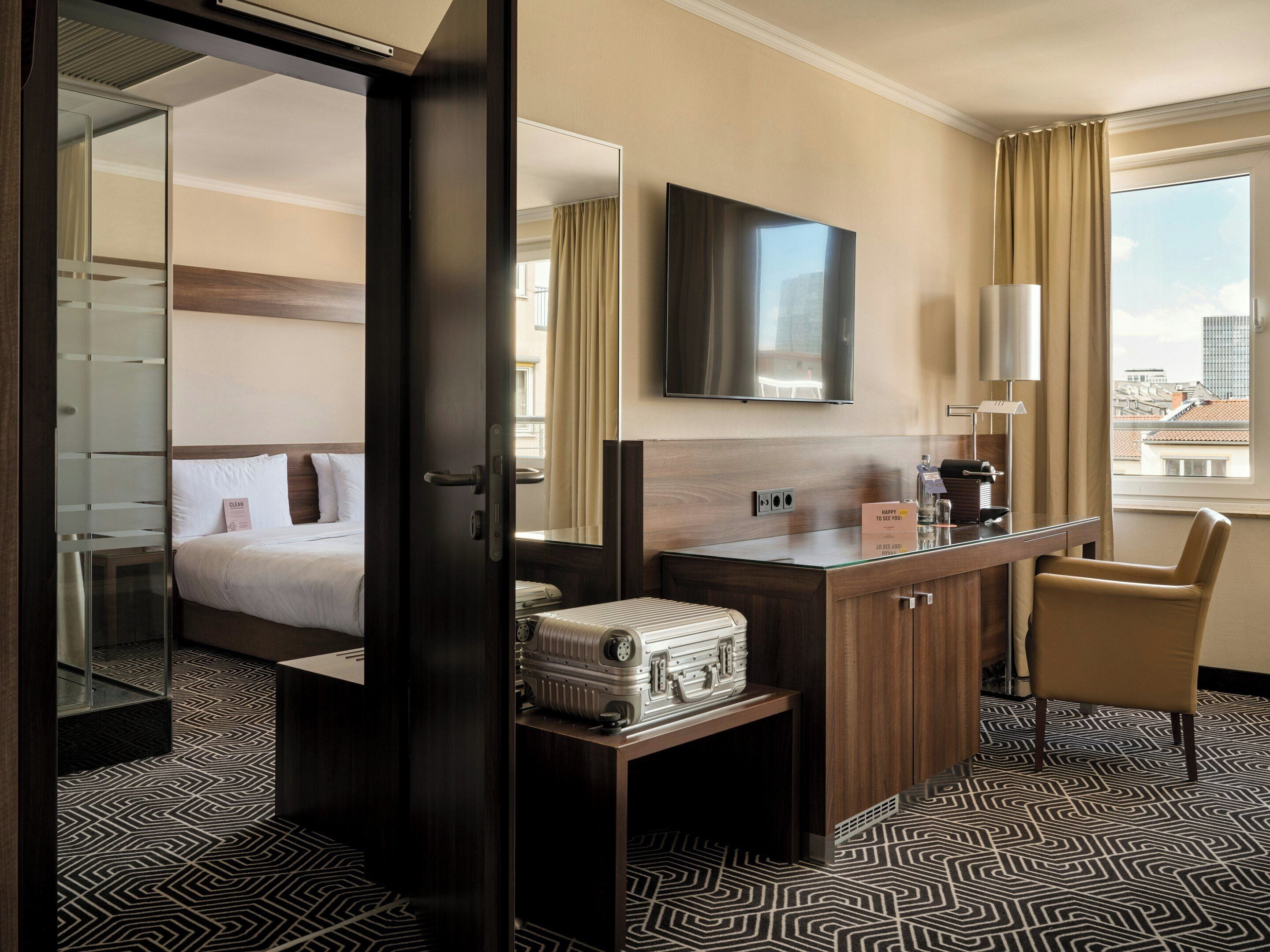 A hotel room with a bed, a desk, a mirror, a television, and a window with curtains.