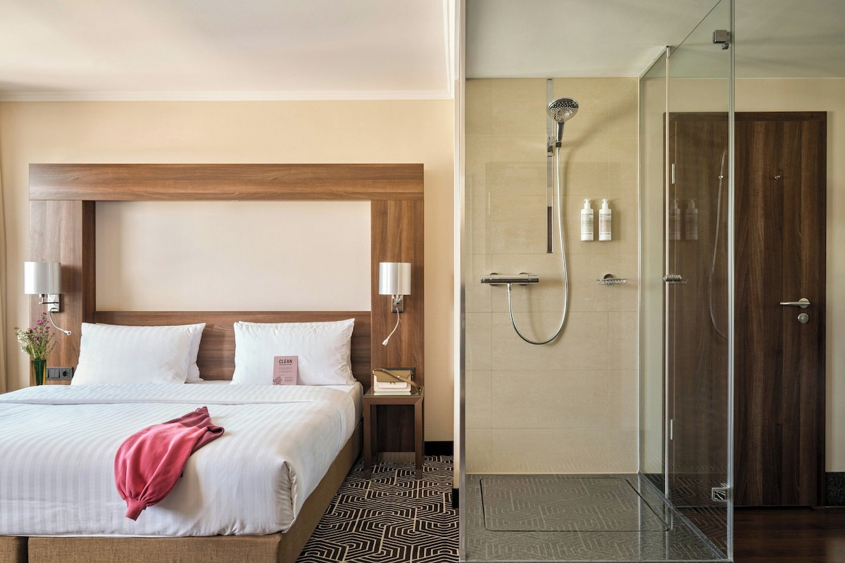 A hotel room with a bed, bedside table, nightstand, and a shower area with a glass door.