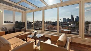 Quarto iluminado pelo sol com vista para a cidade, televisão e assentos confortáveis.
