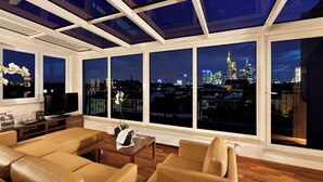 A modern living room with a city view, large windows, and a flat-screen TV. - Flemings Hotel Frankfurt Main-Riverside (Frankfurt)