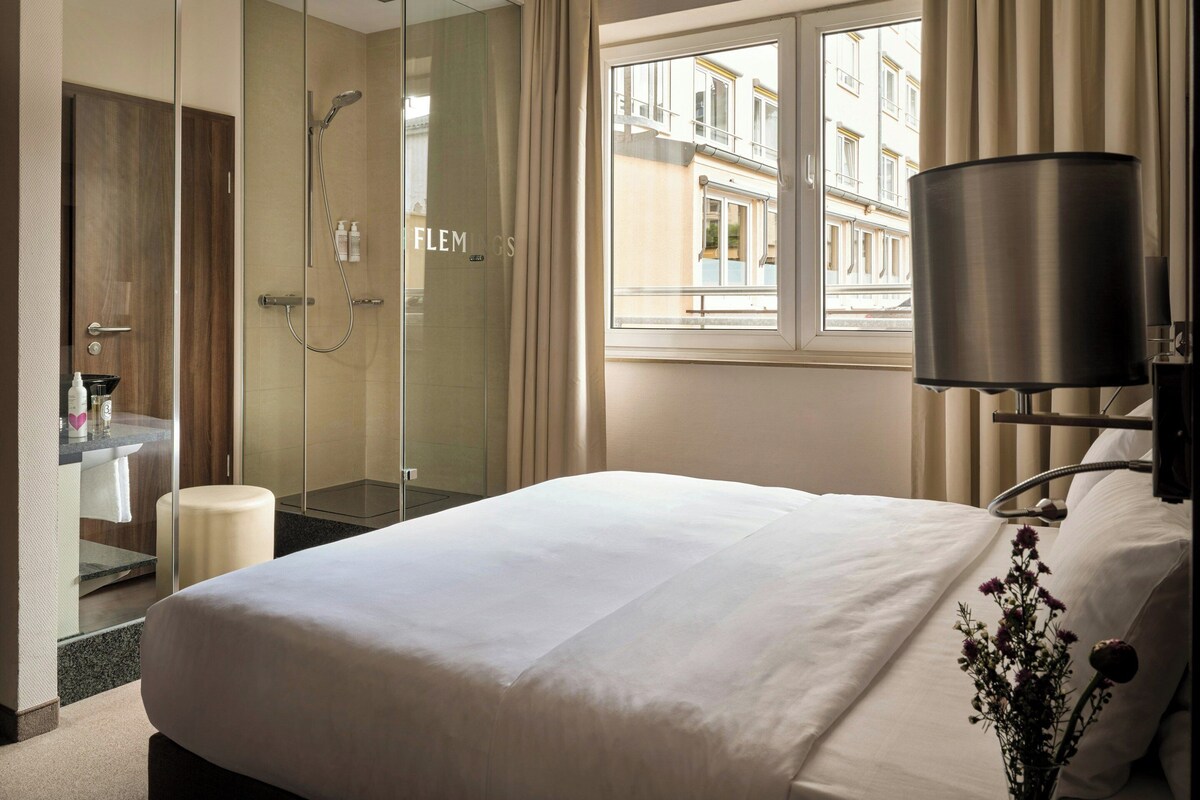 A hotel room with a bed, a glass-enclosed shower, a vanity with a mirror, and a window with a view of buildings.