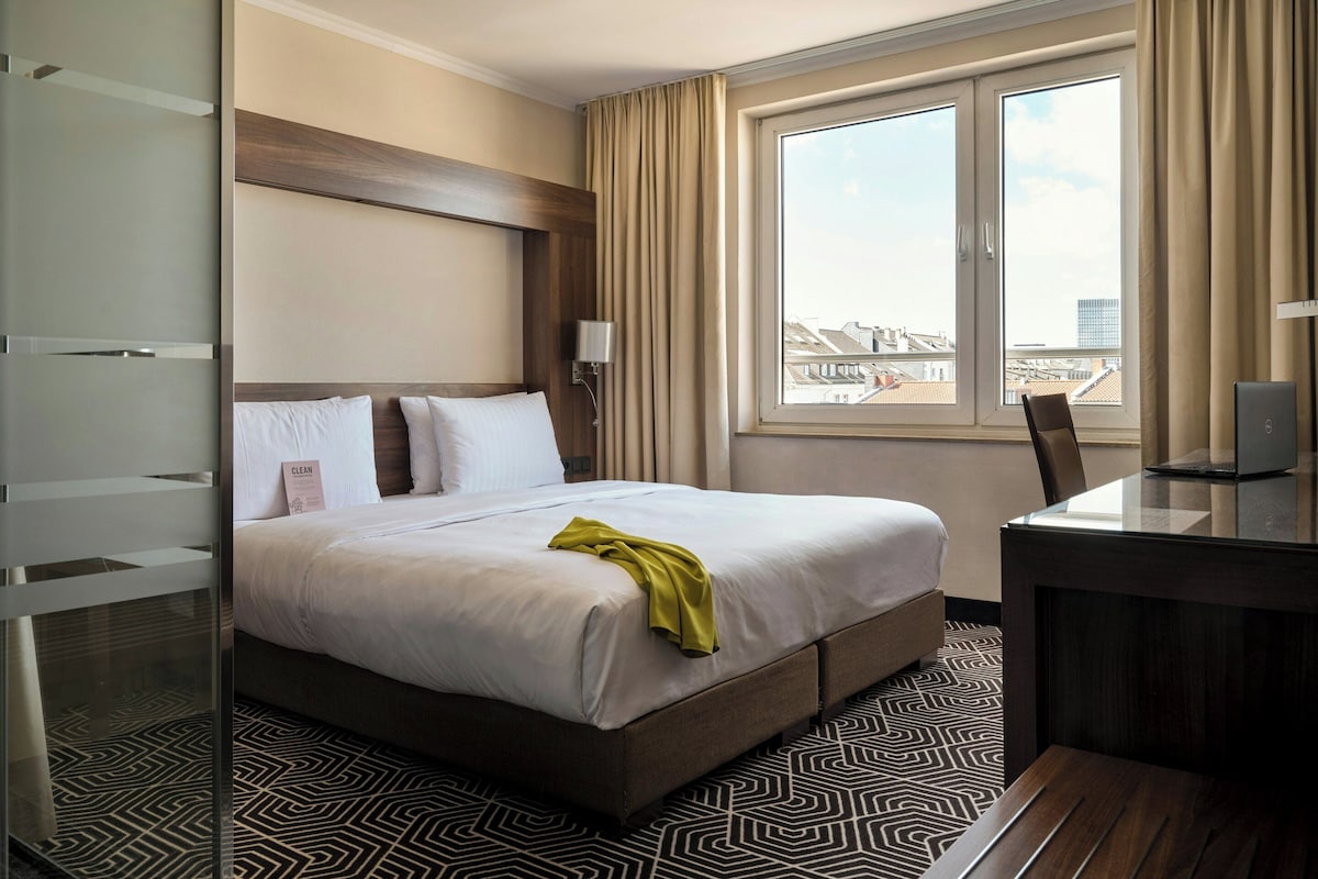 A hotel room with a bed, a desk, a lamp, and a window with a city view.