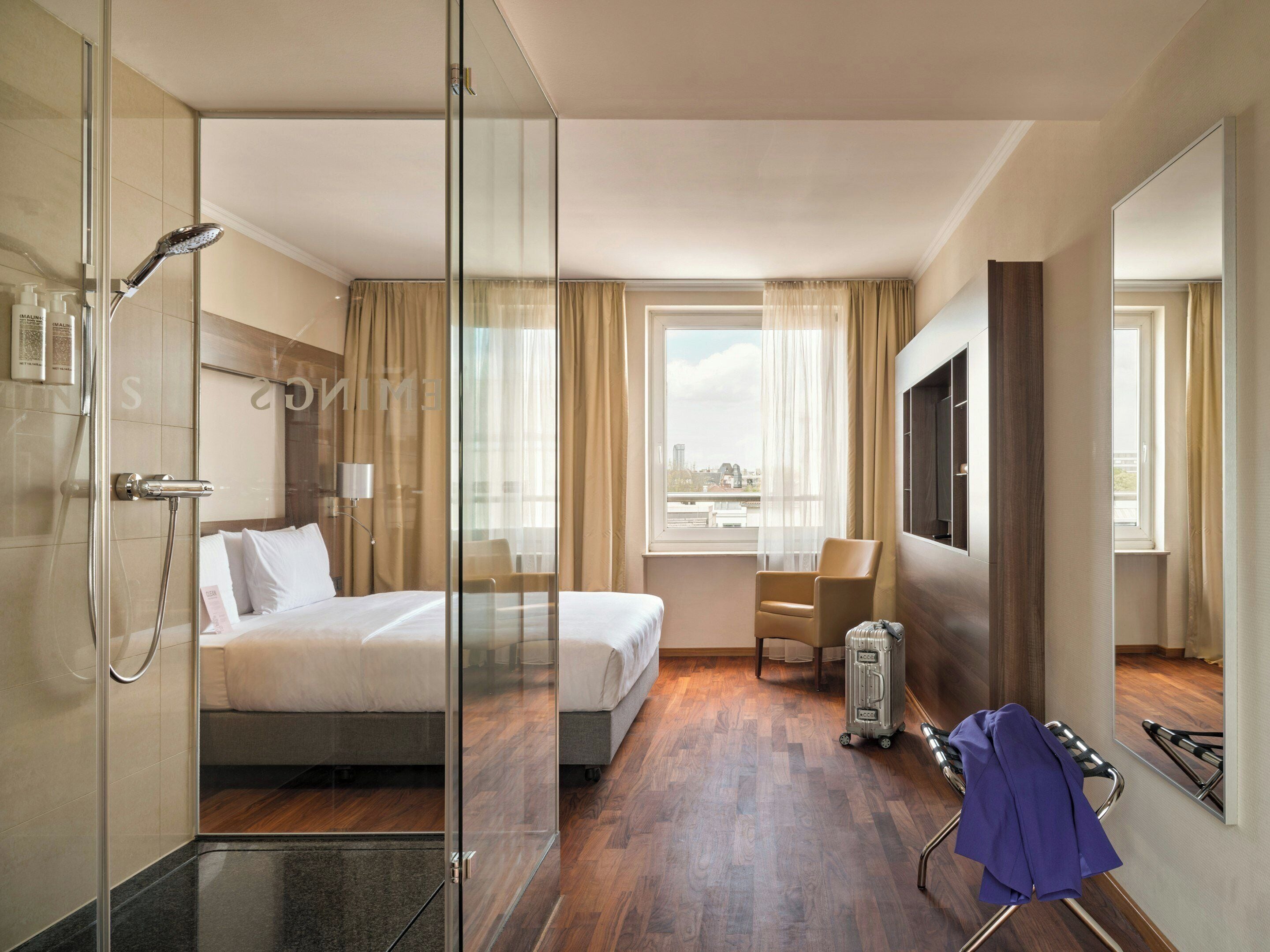 A hotel room with a glass shower enclosure, a bed, a seating area, a TV, and a window with a city view.