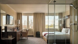 A hotel room with a bed, a desk, a chair, a TV, and a glass-enclosed shower. - Flemings Hotel Frankfurt Main-Riverside (Frankfurt)