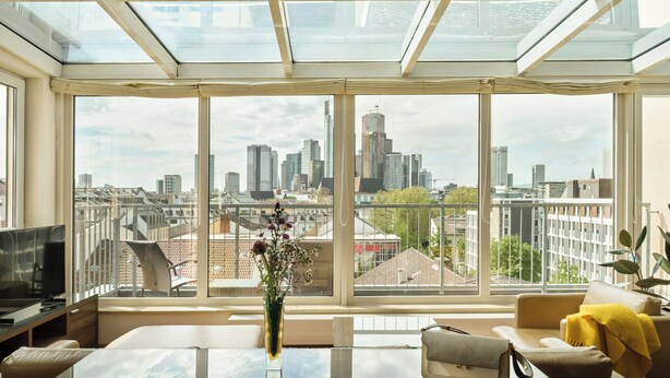 A modern living room with a glass table, a sofa, and a city view through large windows. - Flemings Hotel Frankfurt Main-Riverside (Frankfurt)