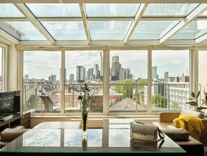 A modern living room with a glass table, a sofa, and a city view through large windows.