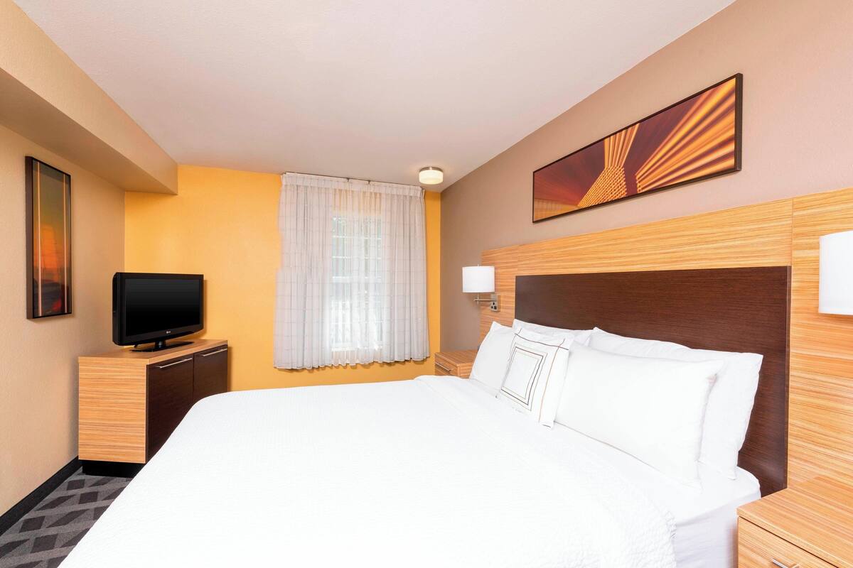 A hotel room with a large bed, a television on a wooden cabinet, a window with curtains, and a wall-mounted picture.