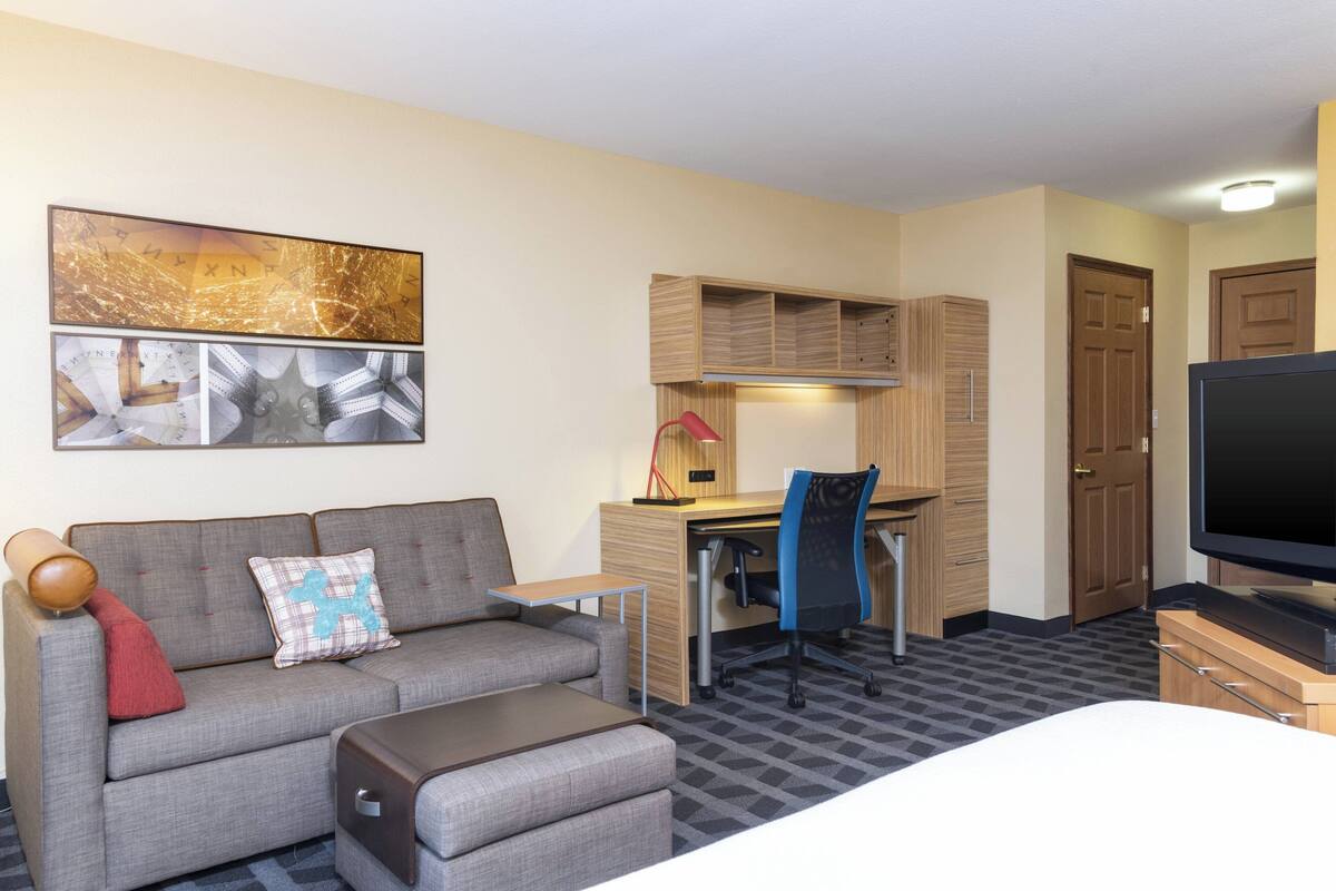A modern hotel room with a grey sofa, a desk with a computer, a TV, and a large abstract painting on the wall.