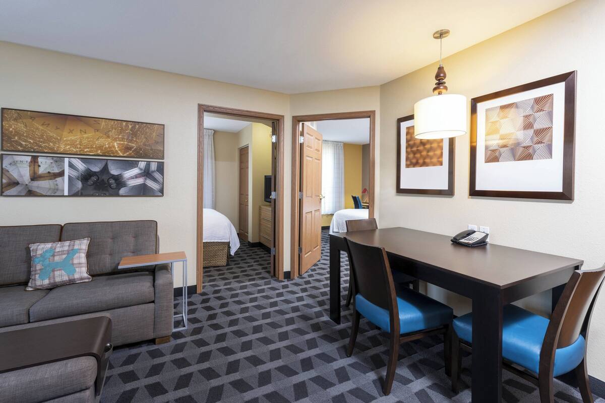A hotel room with a sofa, a small table, a dining table with chairs, and artwork on the walls.