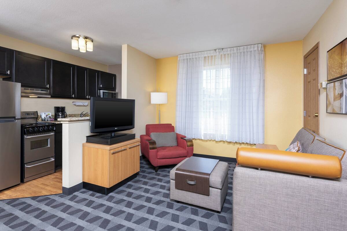 A compact living space with a kitchenette, a flat-screen TV, a red armchair, a sofa, and a patterned rug.