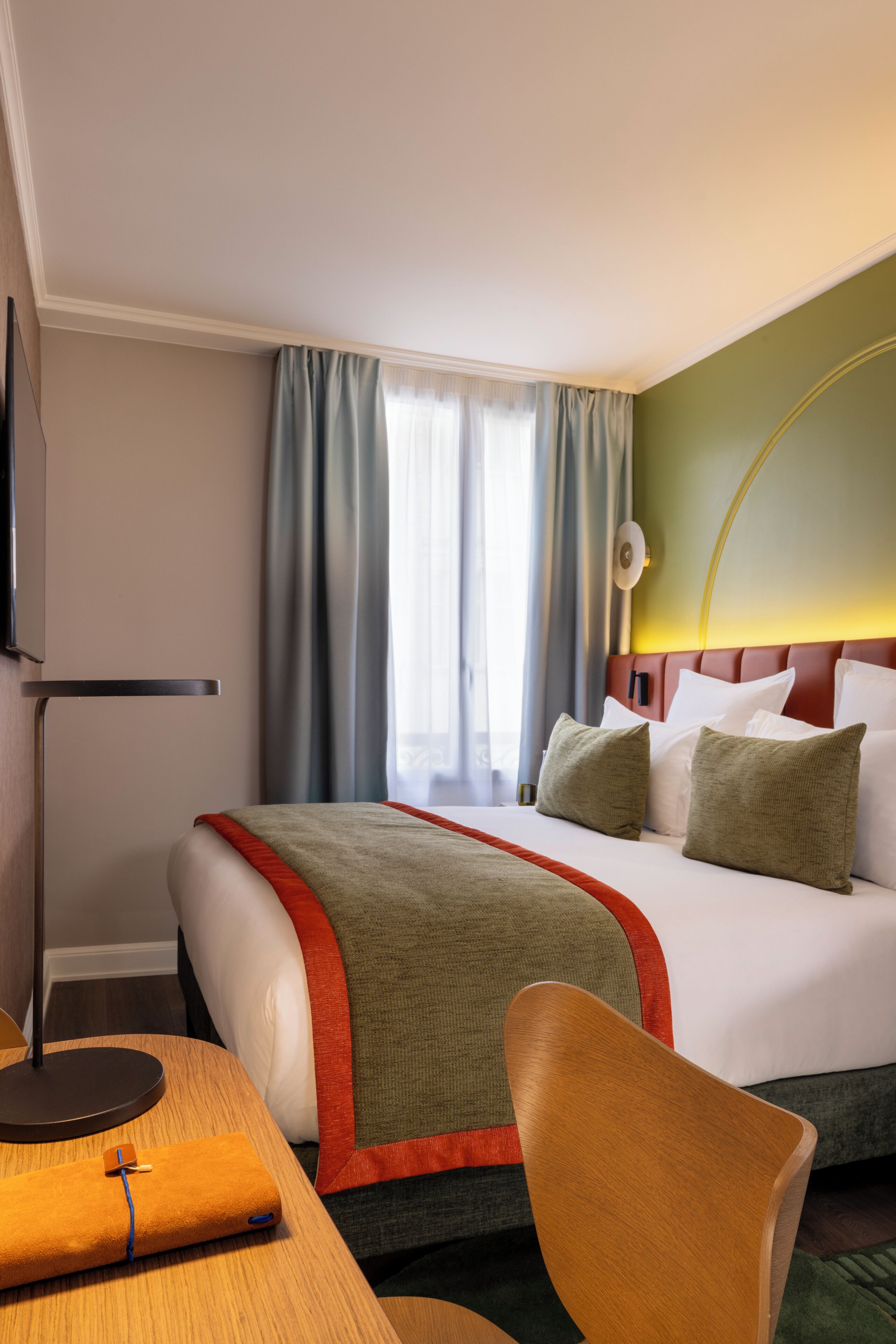 A hotel room with a bed, a desk with a lamp, a chair, and a television on the wall.