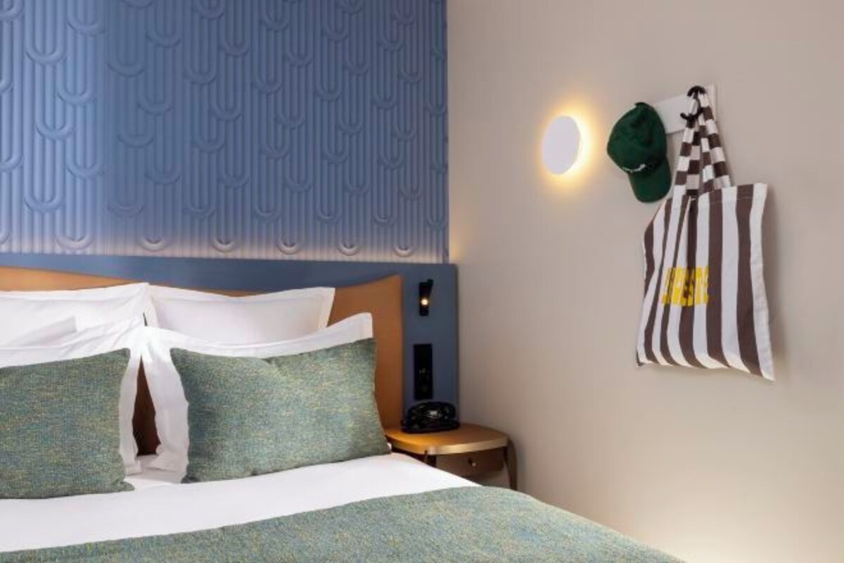 Una cama bien tendida con almohadas, un cabecero con estampado geométrico, una mesita de noche con un teléfono y un perchero de pared con una toalla a rayas.