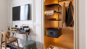 A modern room with a flat-screen TV, a desk with a laptop, a chair, a small ottoman, a shoe rack, and a safe. - Hôtel L'Ormaie & Spa (Paris)