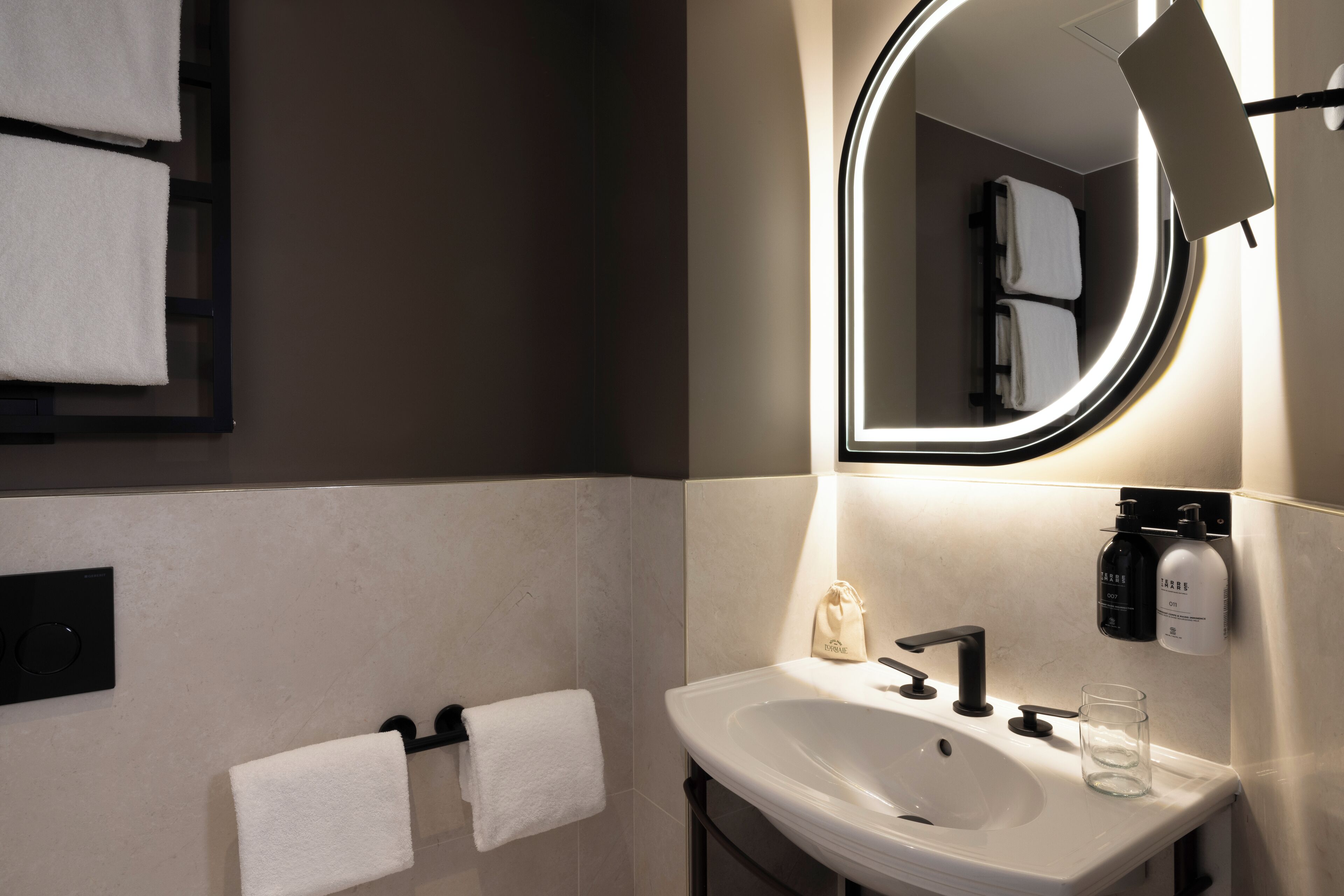 Une salle de bain moderne avec un lavabo blanc, un robinet noir et un miroir rond avec un éclairage intégré. Deux serviettes blanches sont suspendues sur un porte-serviettes, et un verre est posé sur le lavabo.