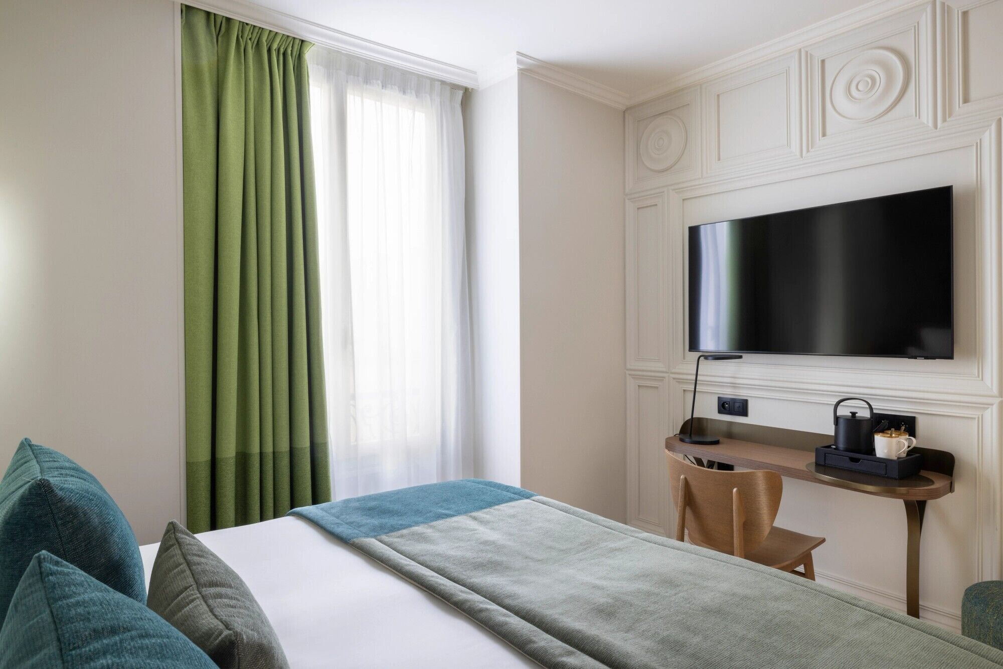 A hotel room with a bed, a desk with a TV, a chair, and a window with green curtains.