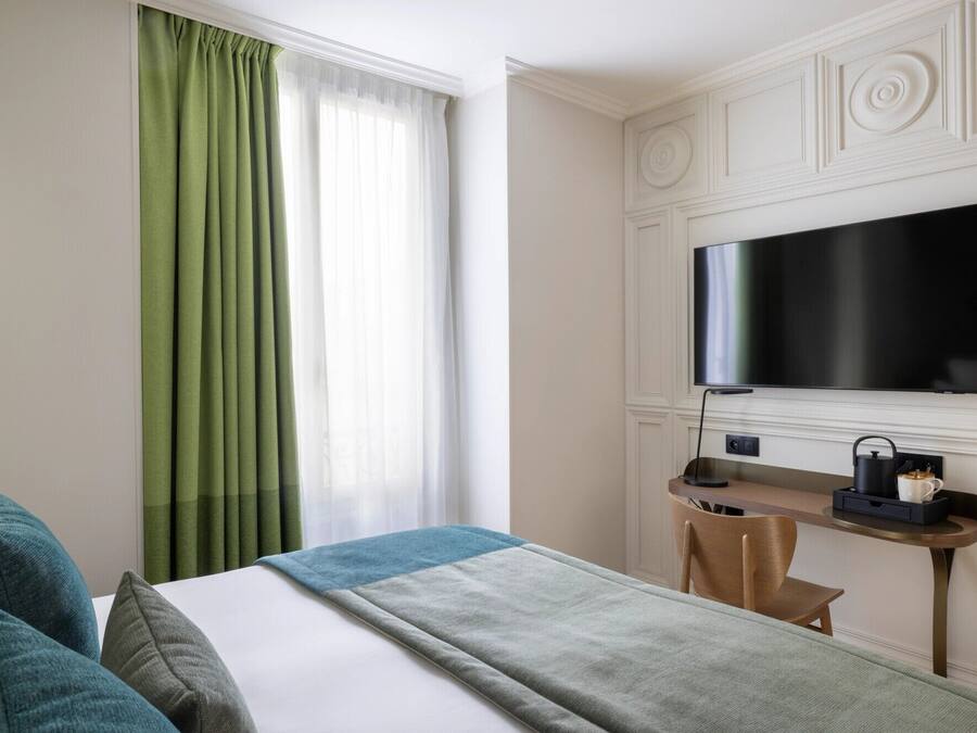 Habitación de hotel con cama, escritorio con televisor, una silla y ventana con cortinas verdes.