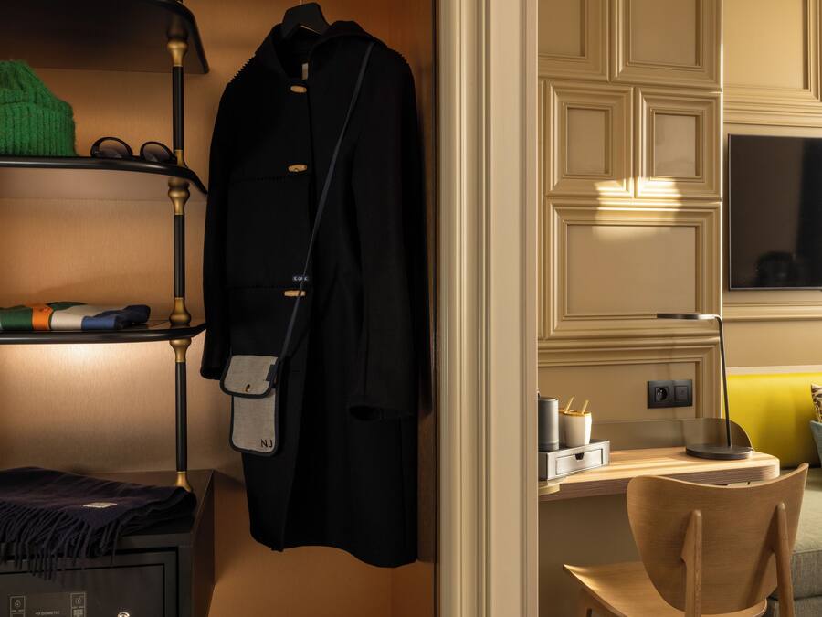 Habitación de hotel con cama, escritorio, silla, televisor, estante con ropa y perchero.