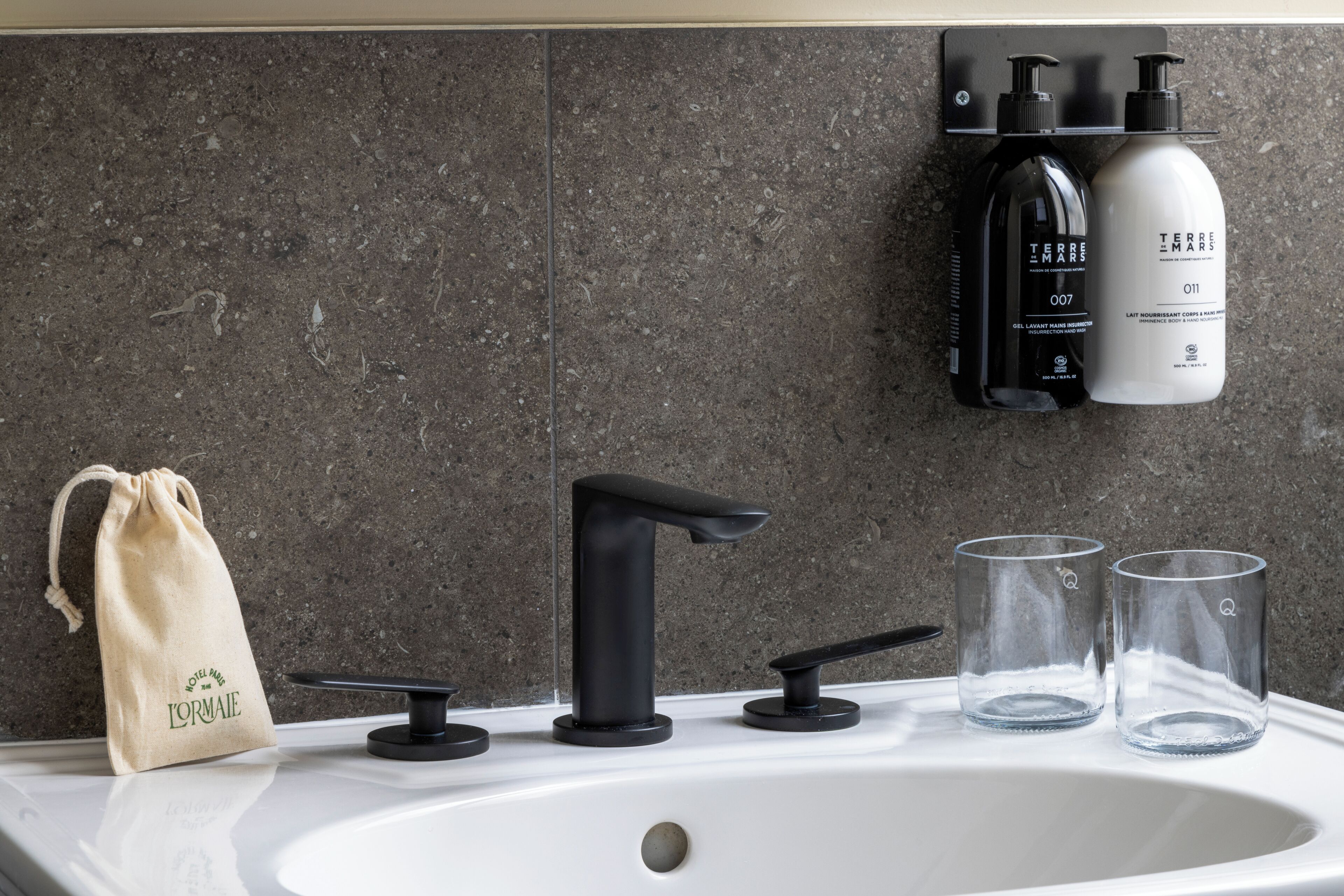Un lavabo de salle de bain avec un robinet moderne, deux verres transparents, un petit sac à cordon et des articles de toilette disposés sur une étagère fixée au mur.