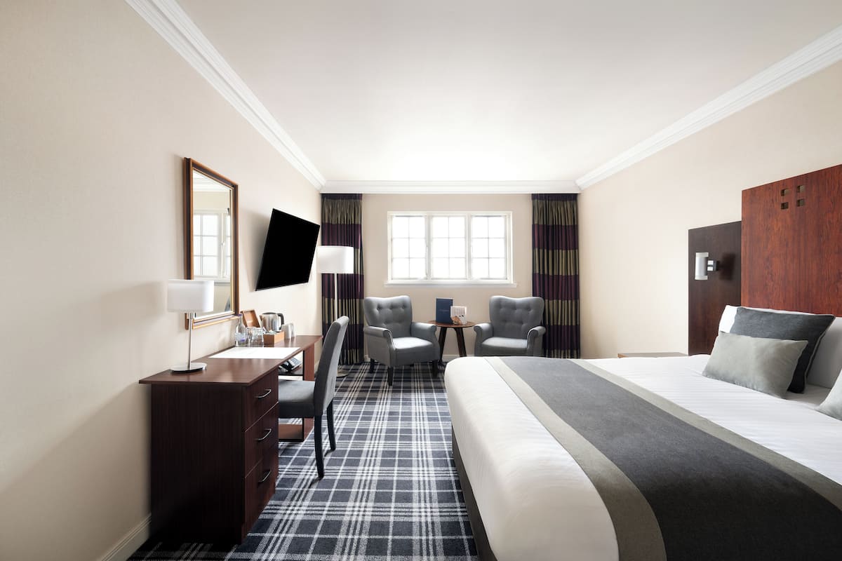 A hotel room with a bed, a desk with a lamp, two armchairs, a TV, and a window with curtains.