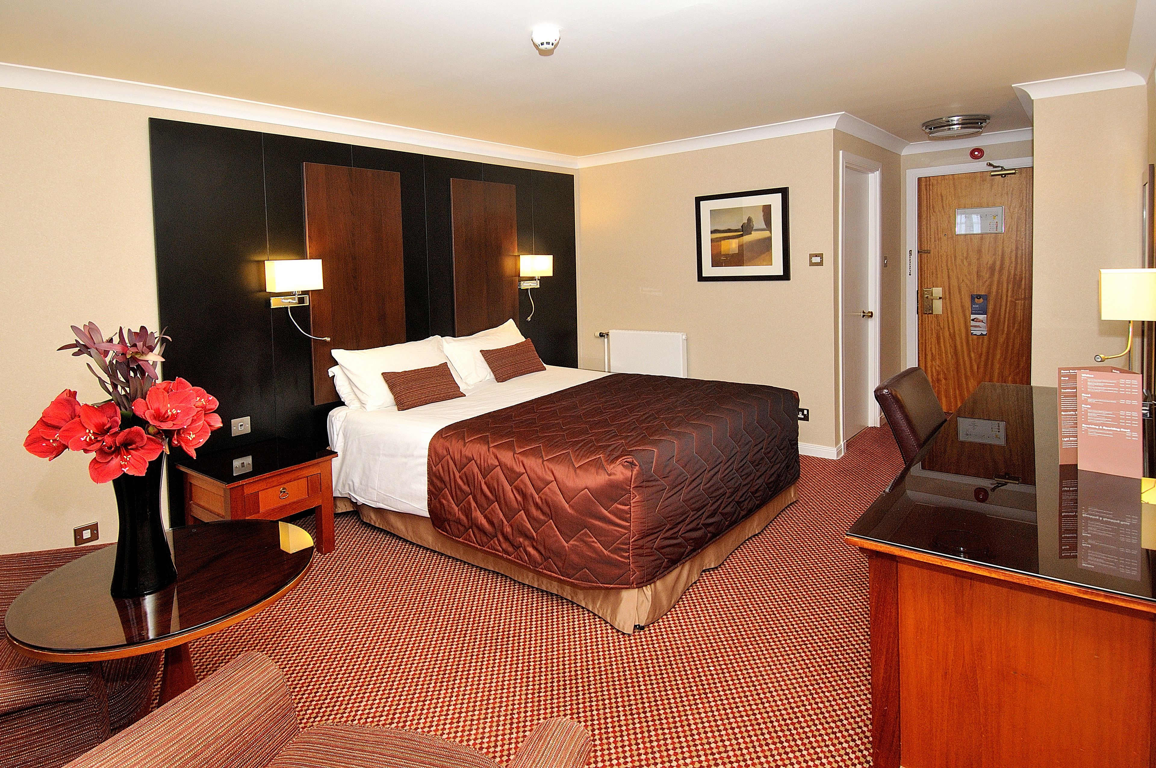 A hotel room with a large bed, a bedside table with a vase of flowers, a desk with a mirror, and a red carpeted floor.