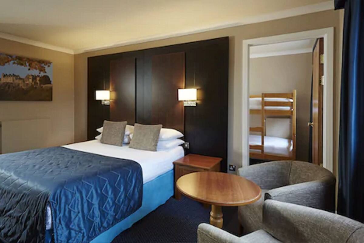 A hotel room with a large bed, two bedside lamps, a small round table, a chair, and a bunk bed in the adjacent room.