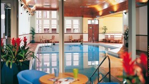 An indoor swimming pool with clear glass walls, wooden ceiling, and seating areas with chairs and tables. - The Stirling Highland Hotel (Stirling)