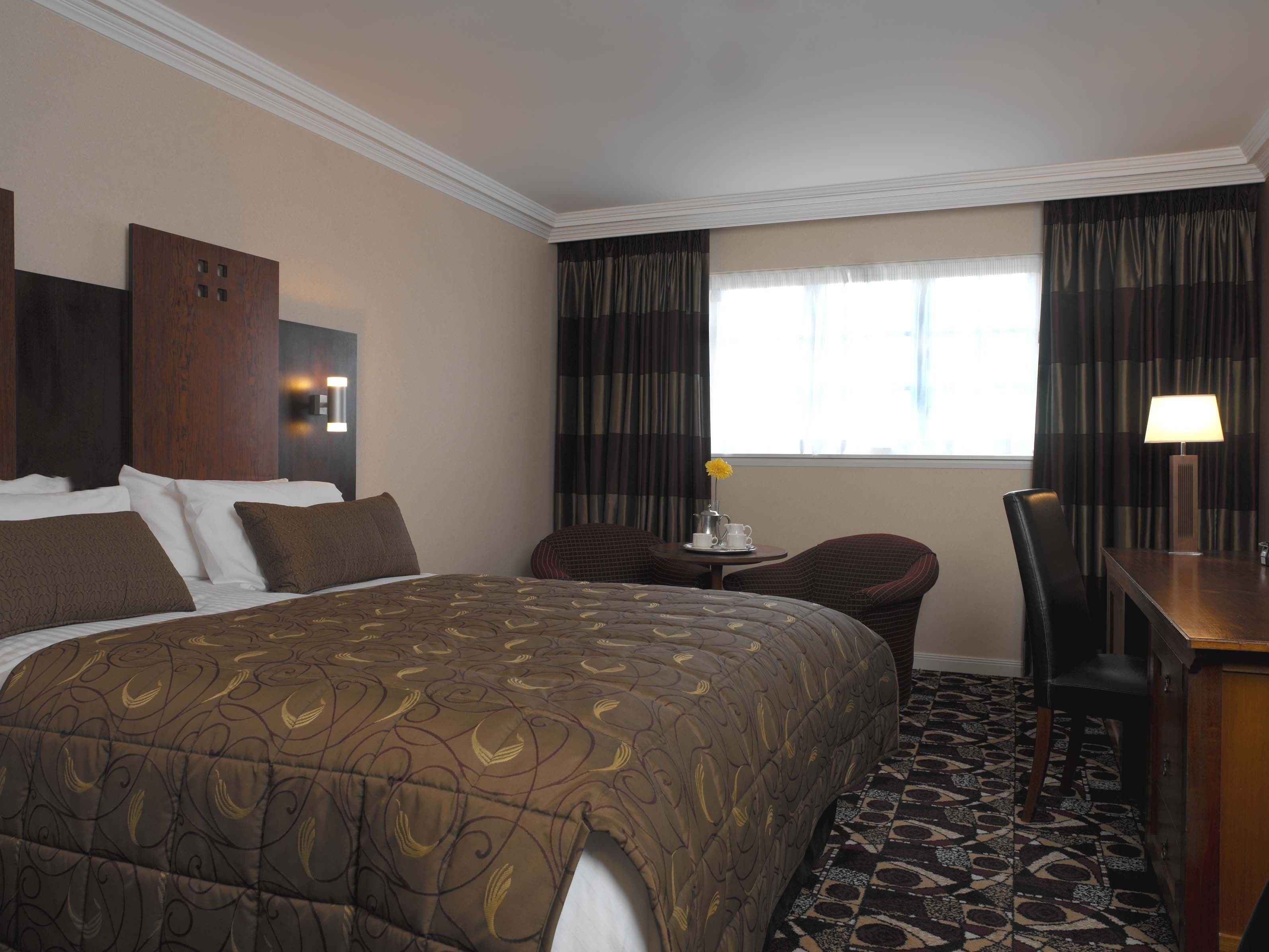 A hotel room with a large bed, a desk, a chair, a small table with a vase and a cup, and a window with curtains.