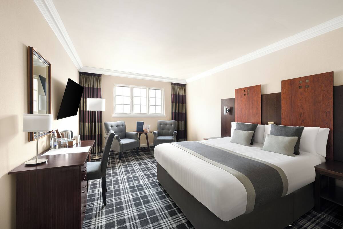 A hotel room with a large bed, two armchairs, a desk with a lamp, a TV, and a window with curtains.
