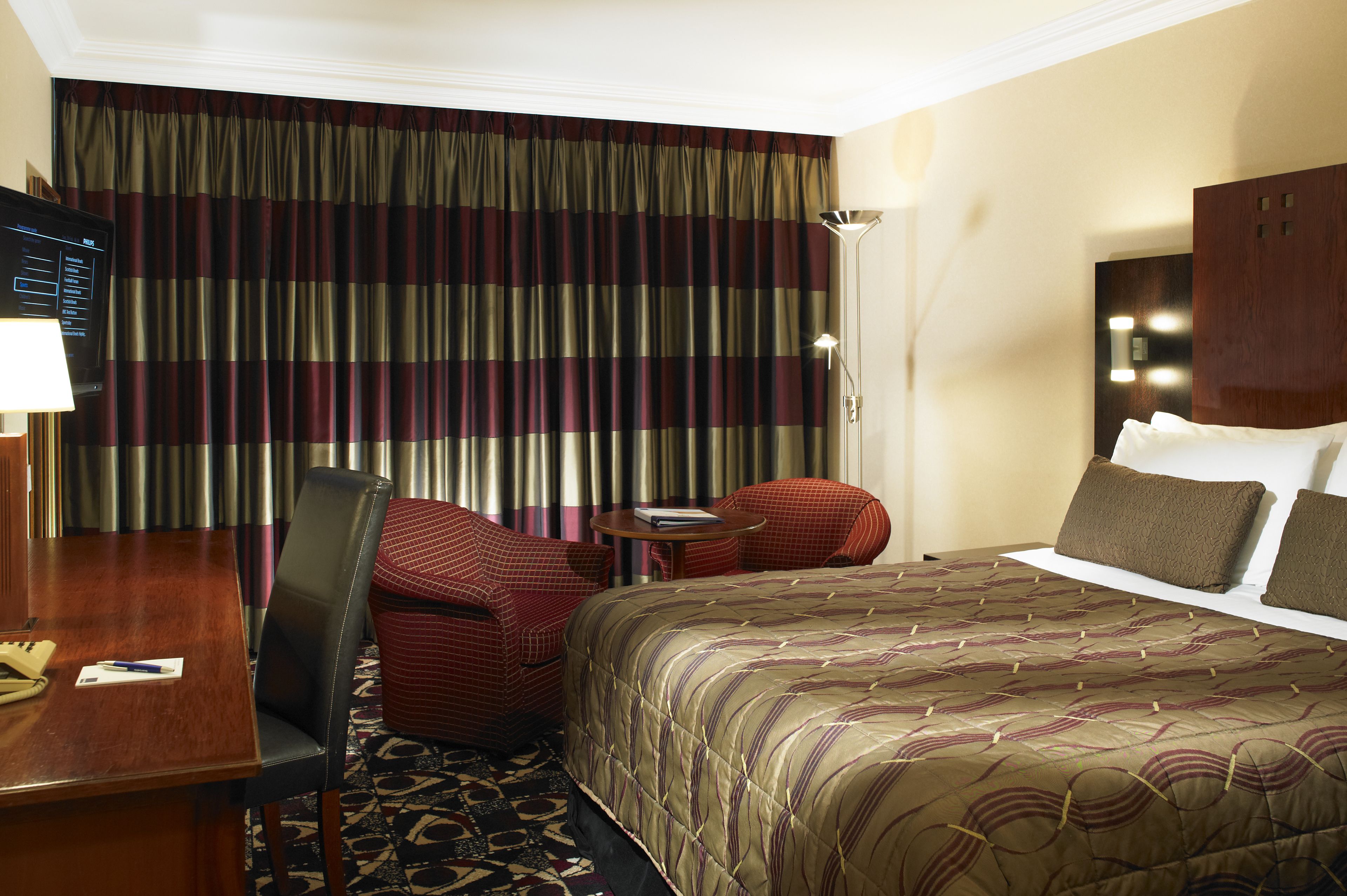 A hotel room with a large bed, a desk with a chair, a small table with a chair, and a large window with curtains.