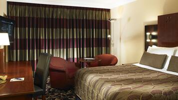 A hotel room with a large bed, a desk with a chair, a small table with a chair, and a large window with curtains.