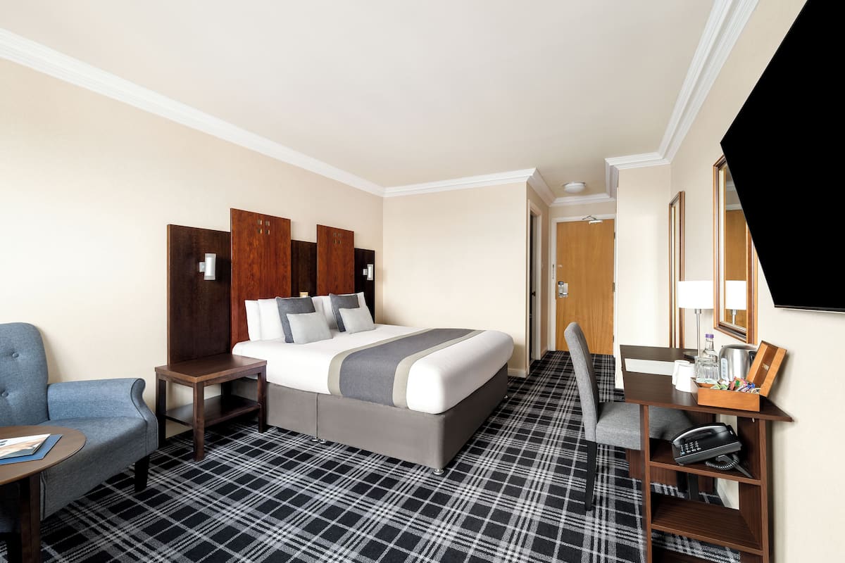 A hotel room with a bed, a desk with a chair, a small table, and a TV.