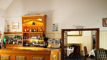 Una zona de bar con mostrador de madera, taburetes y una lámpara de araña. Se observa una alfombra estampada y un comedor con mesa y sillas.