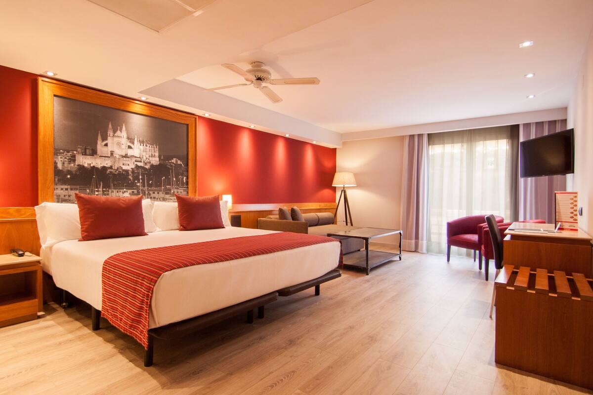 Una habitación de hotel con una cama grande, un escritorio, una silla, una lámpara de escritorio, un televisor y un ventilador de techo.