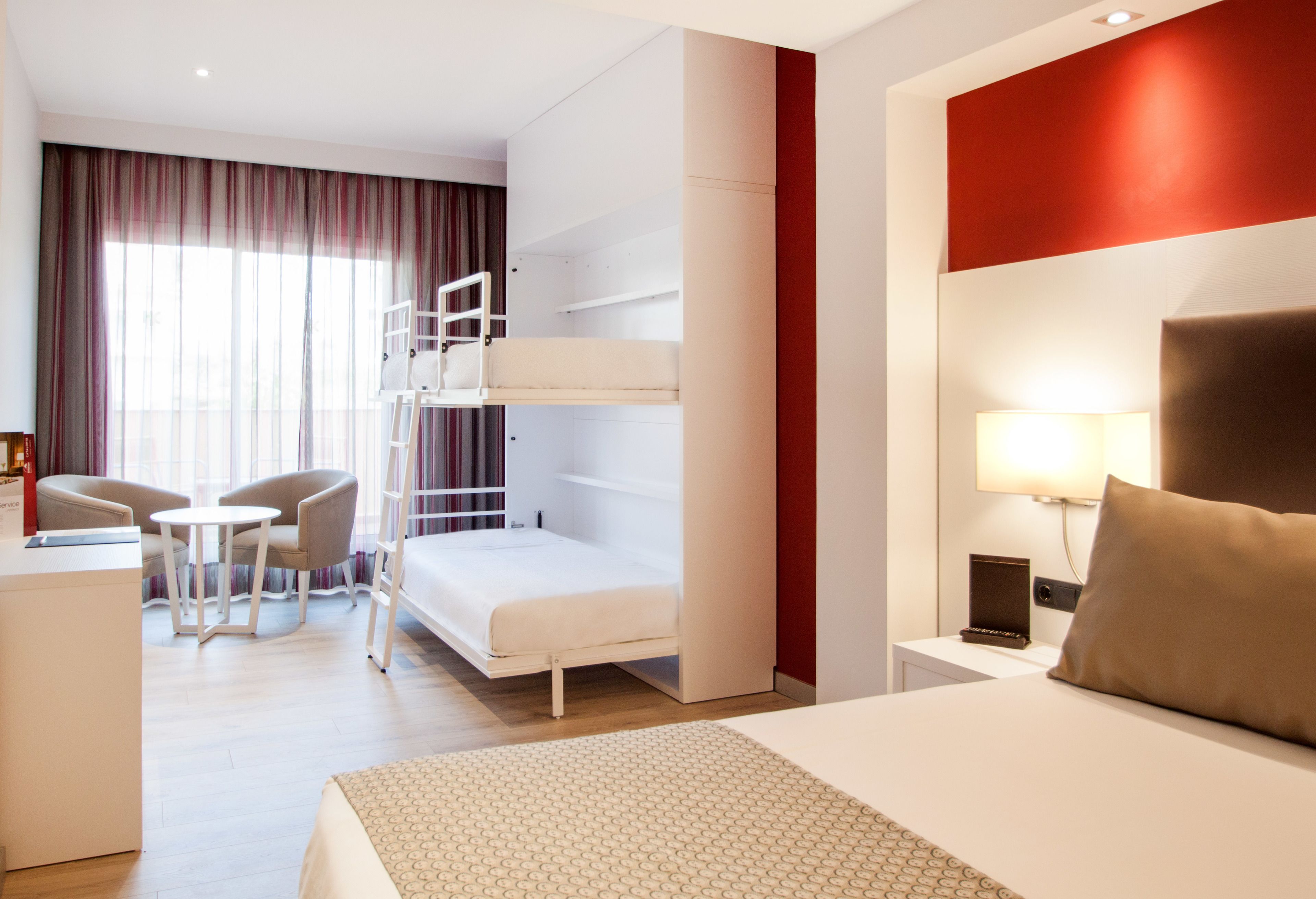 Une chambre d’hôtel avec un lit superposé, un bureau, deux chaises, une table de chevet avec une lampe et un mur d’accent rouge.