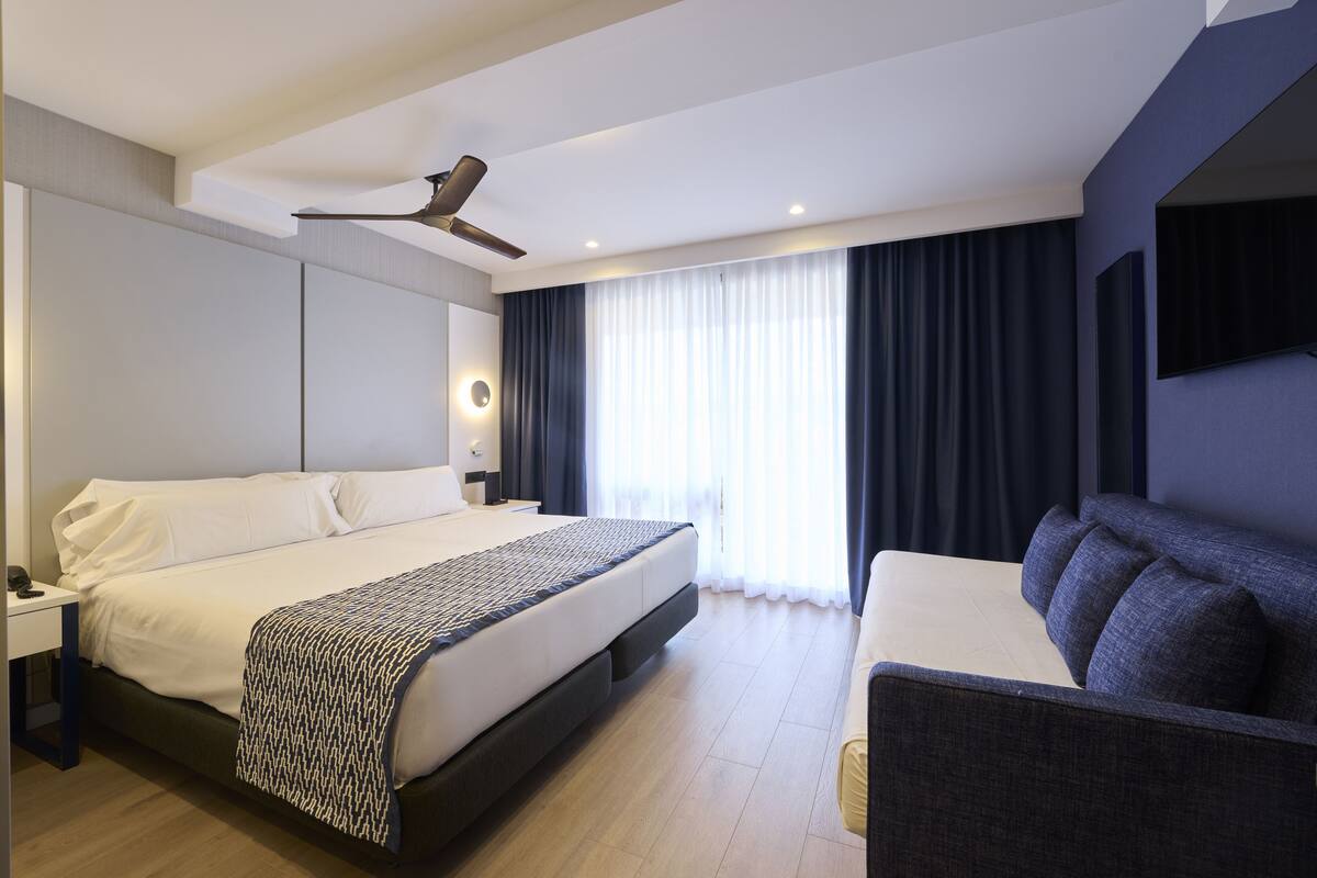 Una habitación de hotel moderna con una cama grande, un sofá, un ventilador de techo y un ventanal con cortinas.