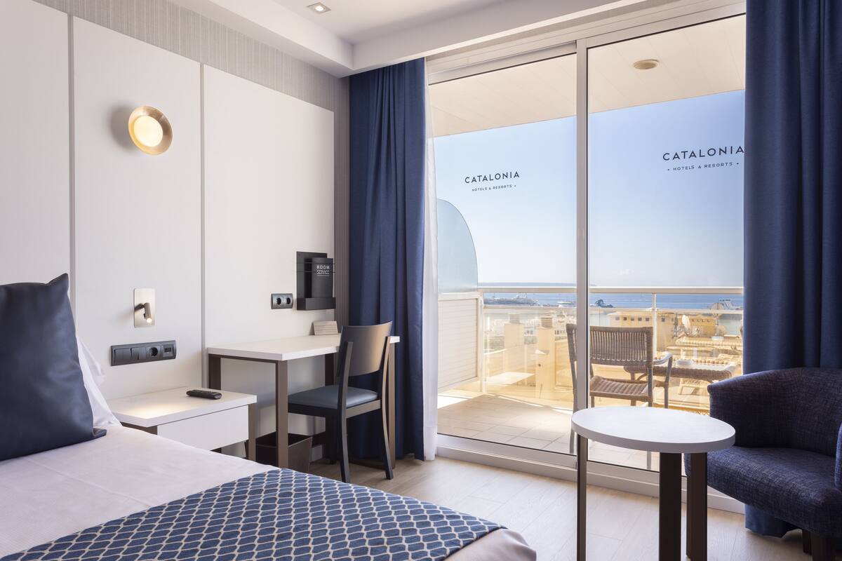 Una habitación de hotel moderna con cama, escritorio, silla y una pequeña mesa. Se ve la ciudad a través de amplios ventanales.