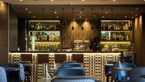 Zona de bar con varias estanterías llenas de botellas de licor, una máquina de café y una barra con caja registradora. Varias mesas y sillas dispuestas para sentarse.
