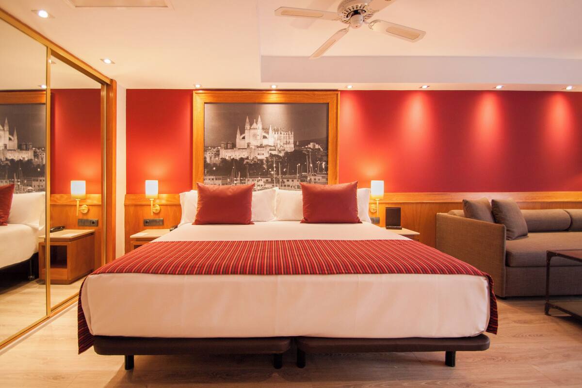 Una habitación de hotel con una cama grande, dos mesitas de noche, un sillón y un ventilador de techo.