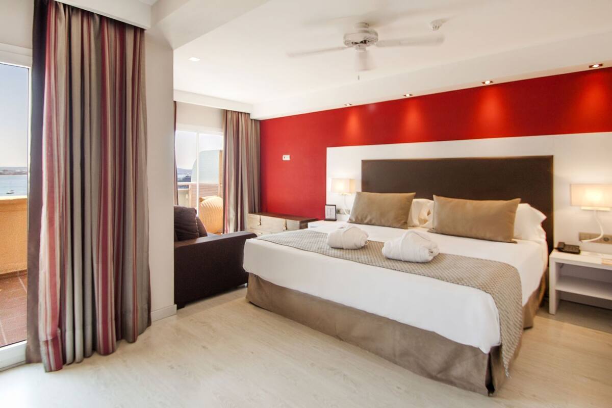 Habitación de hotel con una cama grande, ventilador de techo, vista al mar y balcón con cortinas.