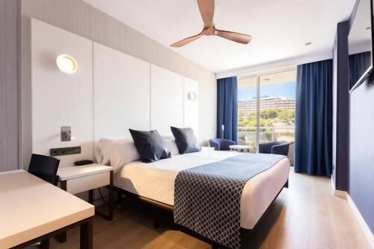 Habitación de hotel con cama, escritorio, silla, ventilador de techo y vista a la ciudad.