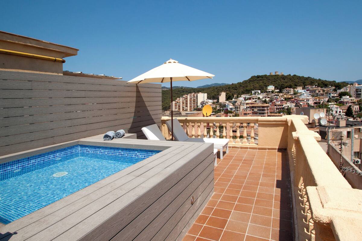 Una piscina en la azotea con cubierta de madera, un paraguas blanco y vista panorámica de una ciudad con edificios y colinas.