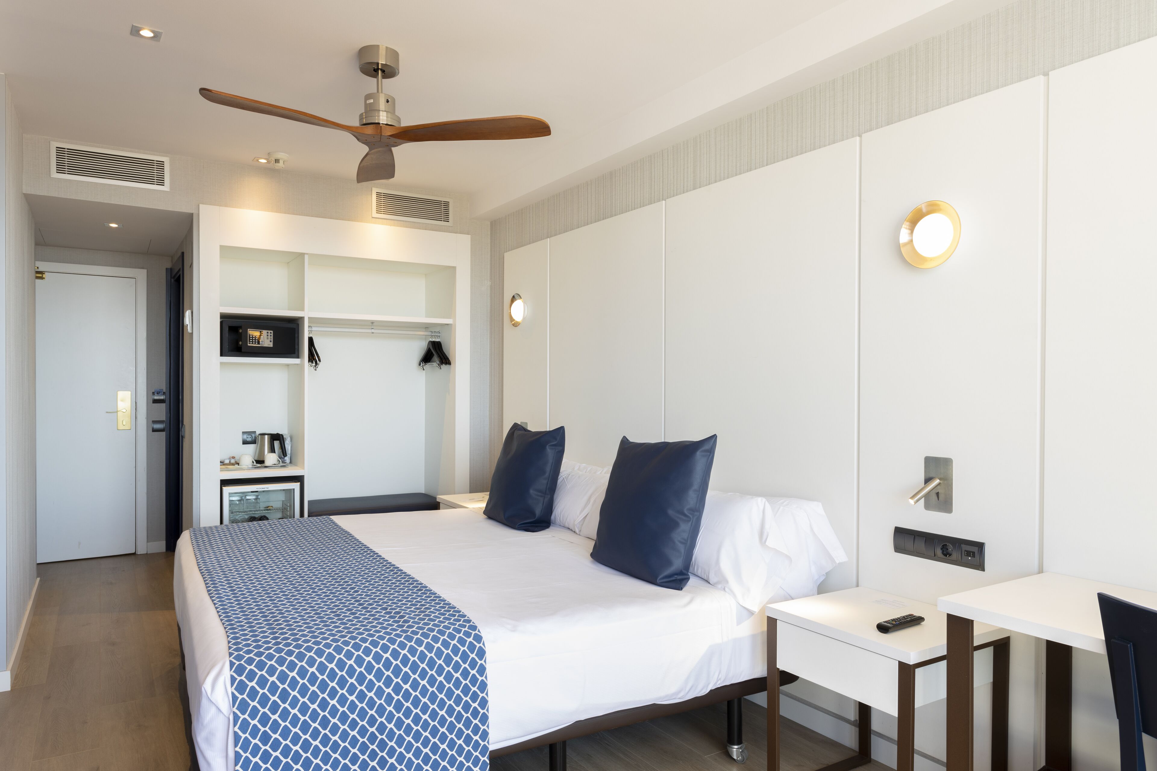 A hotel room with a bed, bedside table, desk, chair, ceiling fan, and built-in shelving.