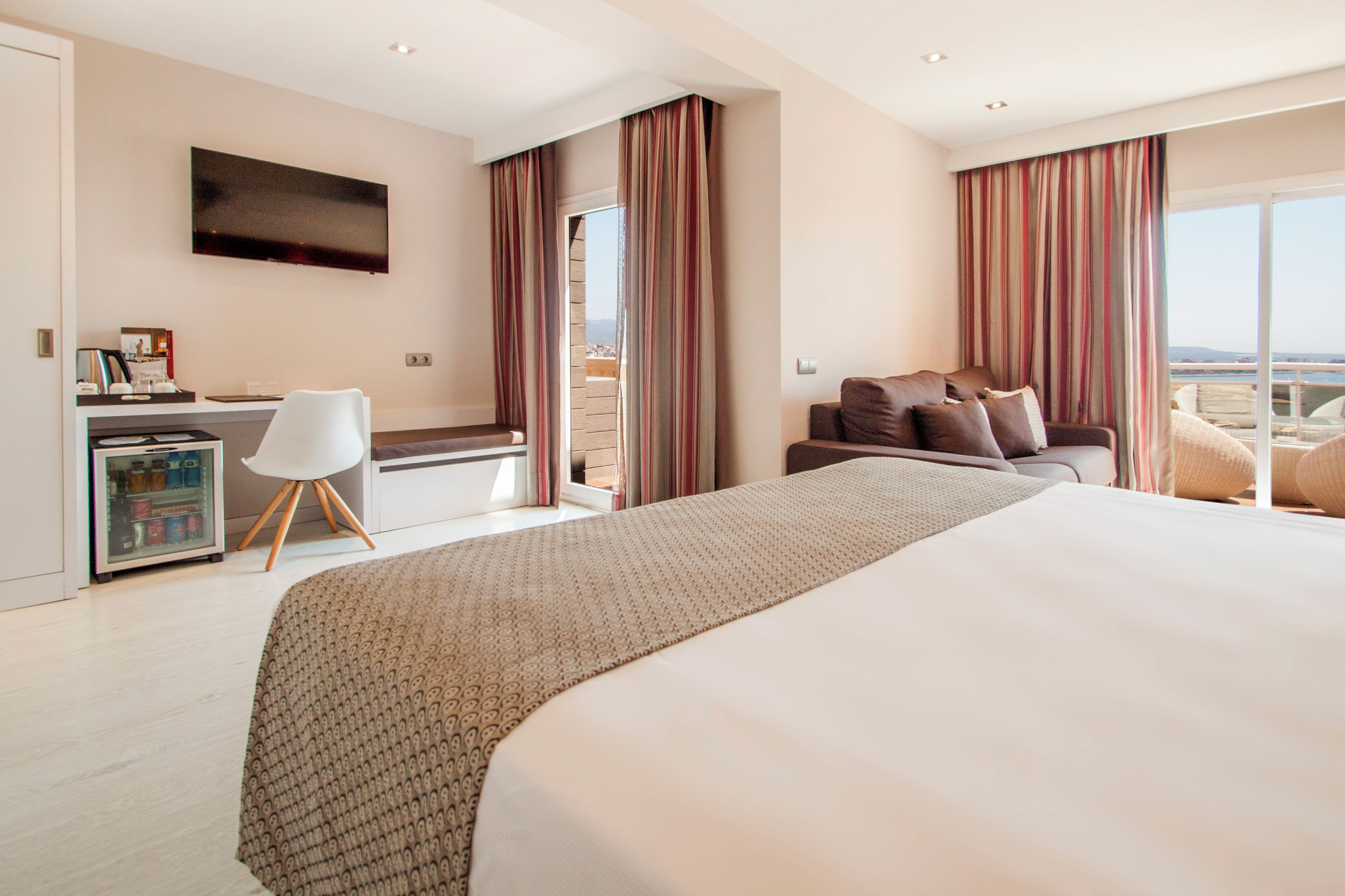 Habitación de hotel con una cama grande, un escritorio con computadora, una silla, televisor y un balcón con cortinas.