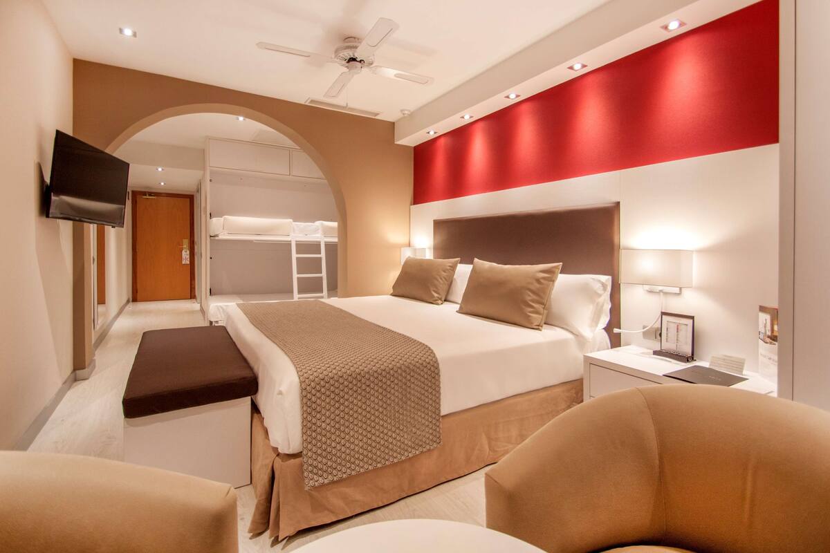 Habitación de hotel con cama, escritorio con lámpara, televisor montado en la pared y ventilador de techo.