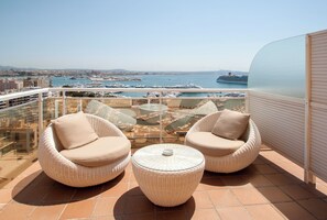 Un balcón con dos sillas de mimbre y una mesa de vidrio, con vista a un puerto deportivo con barcos y un crucero.
