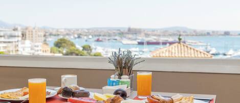 Taula de petit esmorzar amb diferents aliments i begudes, vista d'una marina amb vaixells i un paisatge urbà al fons.