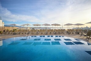 Zona de piscina con sillones de playa y sombrillas, cielo despejado y vista al mar y embarcaciones en el fondo.